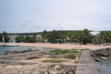 Imagine a panoramic view of Grand Lido Negril's exclusive, clothing-optional beach. The image captures the serene beauty of the Caribbean with its crystal-clear turquoise waters gently lapping against the soft, golden sands. Luxurious lounge chairs and private cabanas dot the beach, offering spots for relaxation and solitude. In the distance, the verdant foliage of Jamaican palm trees sways gently in the breeze, framing the picturesque scene.